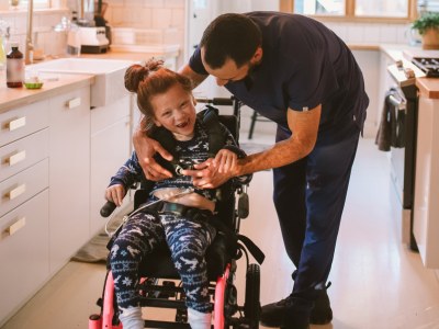 A young girl in a wheelchair laughs joyfully as a caregiver assists her with a medical device.